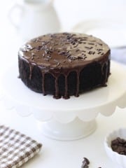 A round chocolate cake with glossy chocolate ganache made from an easy chocolate ganache recipe and chocolate sprinkles sits on a white cake stand. A patterned napkin and a small bowl of chocolate chips are nearby on a white surface.