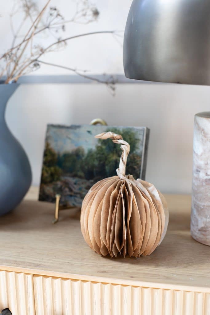A small paper pumpkin decoration sits on a wooden surface, offering inspiration for pumpkin decorating without carving, next to a blue vase with dried flowers, a small framed landscape painting, and a marbled cylindrical container.