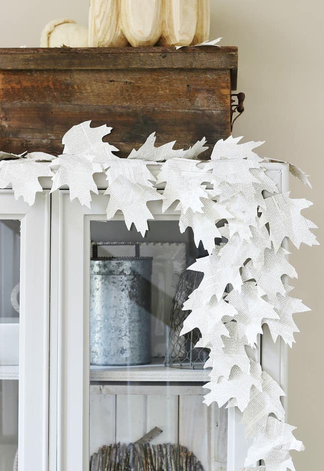 A white cabinet is decorated with my favorite Thanksgiving decor—a garland made of paper leaves cut from book pages, draped along the top edge. A rustic wooden box and a white pumpkin sit on top of the cabinet.