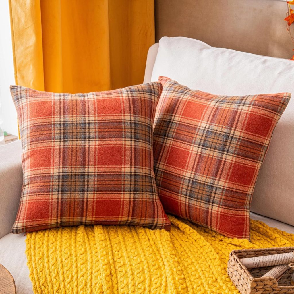 Two plaid pillows with orange, red, and brown tones rest on a white sofa draped with a textured yellow blanket—some of my favorite Thanksgiving decor. A woven basket peeks from the corner while yellow curtains hang in the background.