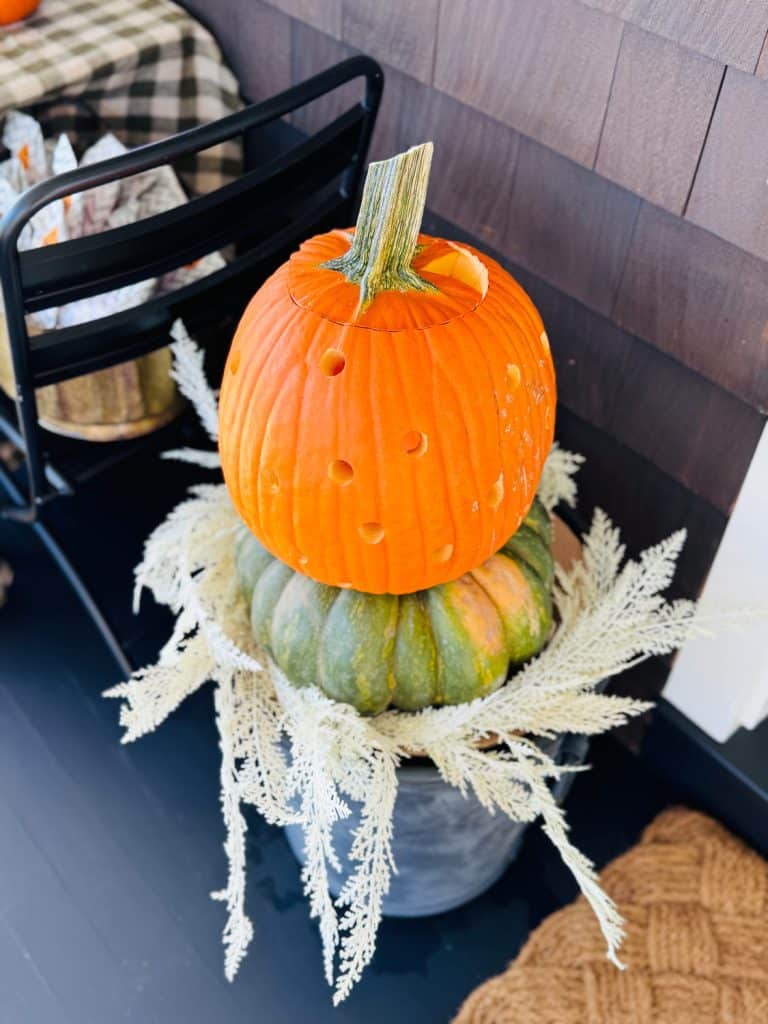 Two stacked pumpkins sit in a pot, topped with white decorative leaves. For creative pumpkin decorating ideas, the top orange pumpkin features small round holes, while the bottom pumpkin is green. A black chair and plaid cloth are in the background.