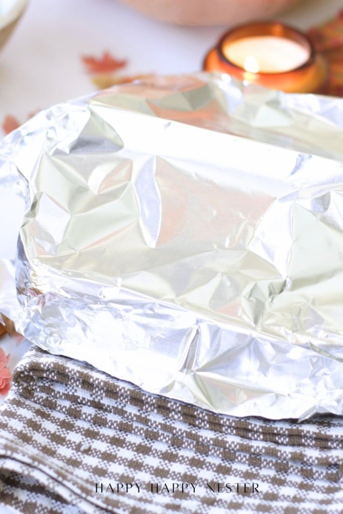 A rectangular dish covered with aluminum foil sits on top of a brown and white checkered cloth, with a lit candle and autumn leaves blurred in the background.