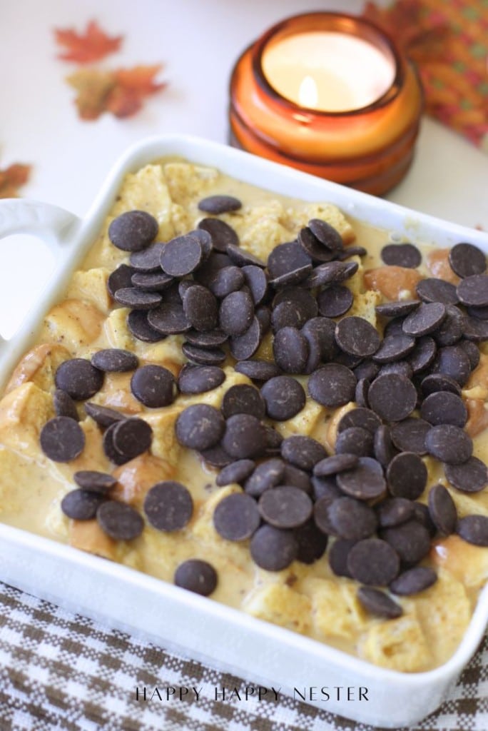 A square white baking dish filled with a creamy dessert topped with chocolate chips sits on a checkered cloth. In the background, there is a lit candle and autumn-themed decorations.