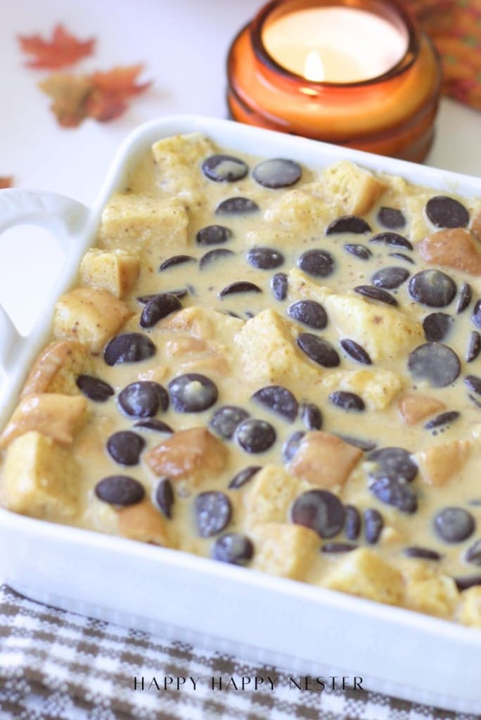 A white ceramic dish filled with pumpkin bread pudding, topped with chocolate chips and apple chunks, with a lit candle in the background.