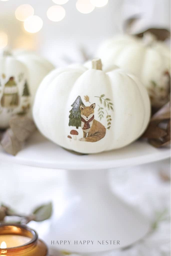 A white pumpkin sits on a cake stand, decorated with a painted fox wearing a scarf, trees, and a mushroom—showcasing pumpkin decorating ideas without carving using temporary tattoos. Soft lights and autumn leaves create a cozy, festive background.