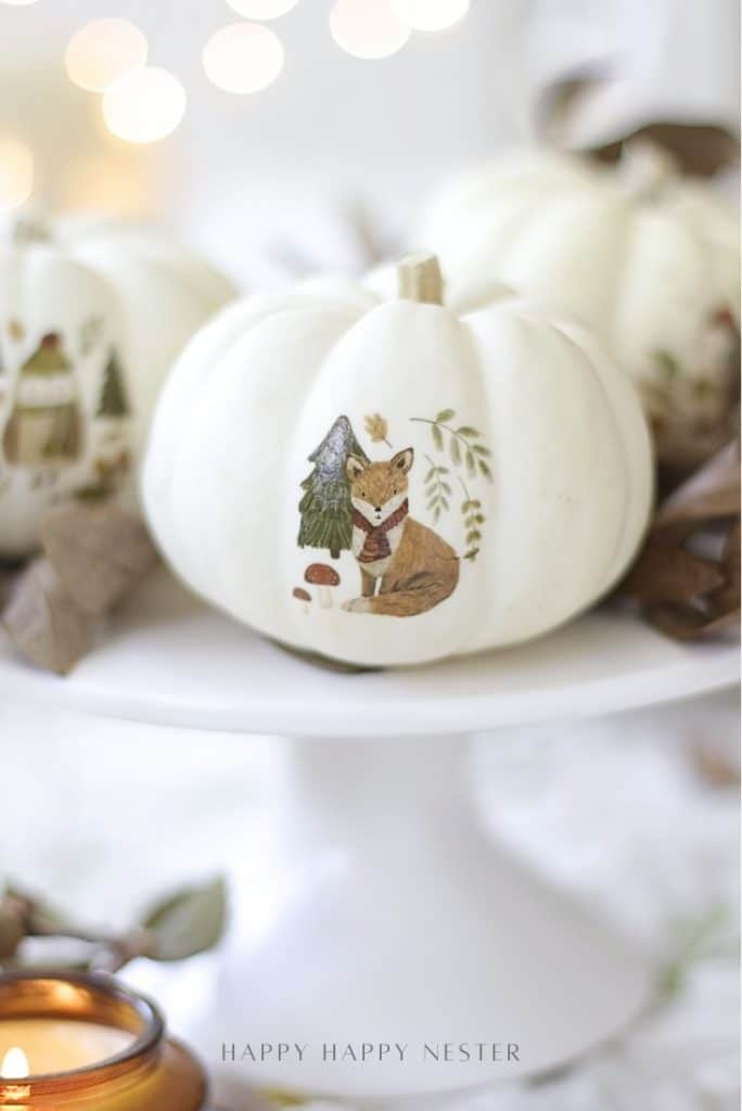 A white pumpkin decorated with a painted fox wearing a scarf, sitting beside a tree and mushroom, is displayed on a white cake stand with autumn leaves and a candle nearby—perfect for Pumpkin Decorating Ideas without carving.