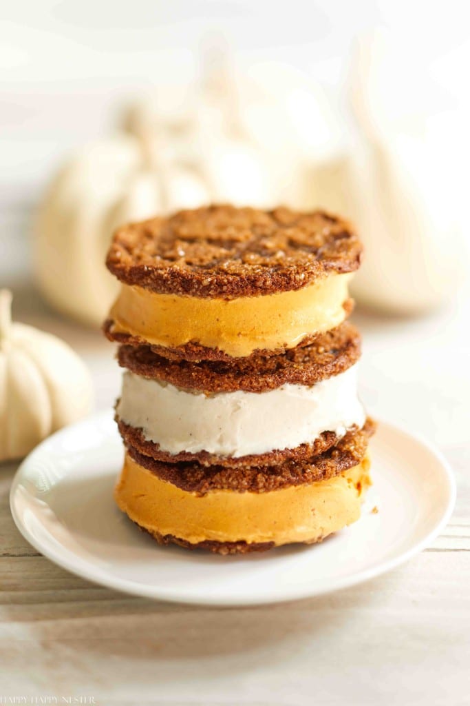 Three ice cream sandwiches made with pumpkin cookies and orange and white ice cream are stacked on a white plate, making for festive Halloween treats with white pumpkins blurred in the background.