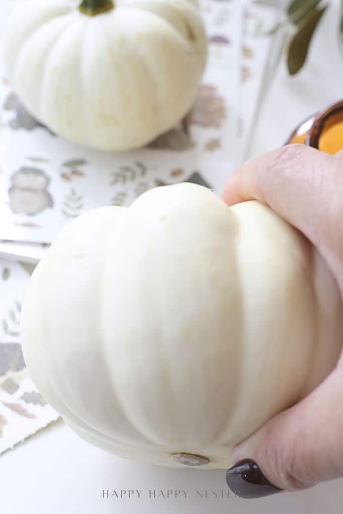 A hand with dark polished nails holds a small white pumpkin; another white pumpkin and decorative papers are blurred in the background.