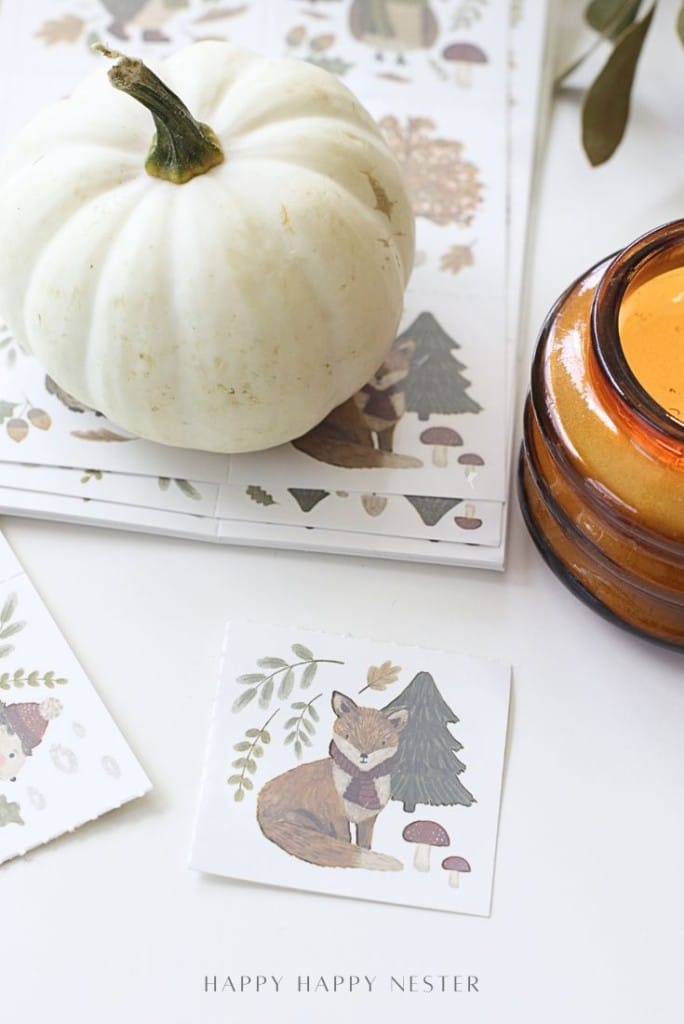 A white pumpkin and an amber candle sit on a table beside woodland-themed stickers, including one featuring a fox wearing a scarf surrounded by leaves, mushrooms, and a pine tree.
