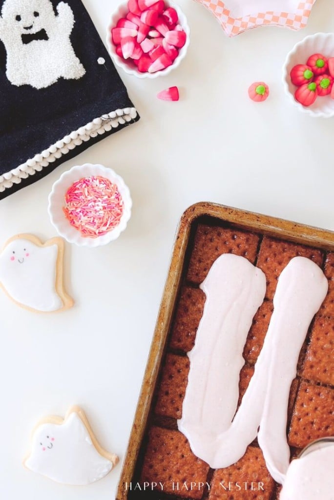 A baking tray of brownies with pink icing beside decorated ghost cookies, a bowl of pink sprinkles, small bowls of candy, and a black napkin with a white ghost design on a white surface.