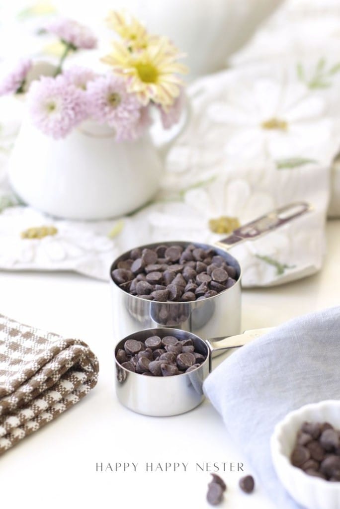 Two metal measuring cups filled with chocolate chips sit on a white surface, ready for your easy chocolate ganache recipe, surrounded by folded napkins, a small bowl, and a white vase with light purple and yellow flowers.