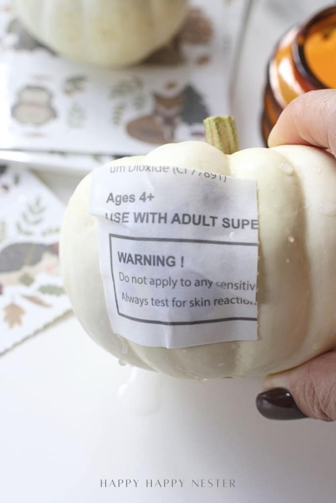 A hand holds a small white pumpkin with a damp warning label that reads "Ages 4+. Use with adult supervision. WARNING! Do not apply to any sensitive areas. Always test for skin reaction.