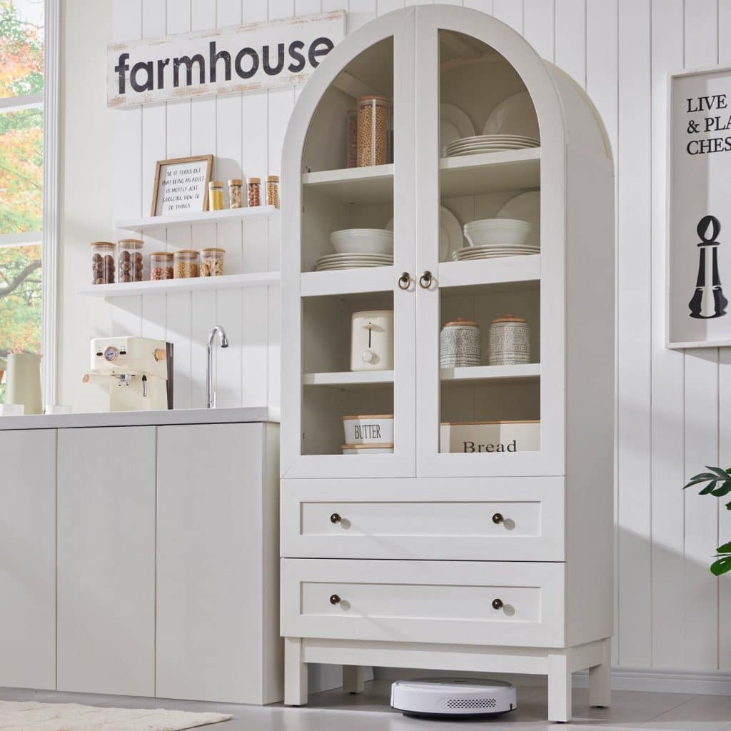 A white arched cabinet with glass doors displays dishes and pantry items; two drawers below. The cabinet stands beside a white kitchen counter, above which hangs a “farmhouse” sign and charming wall crafts for fall. A robot vacuum sits beneath the cabinet.