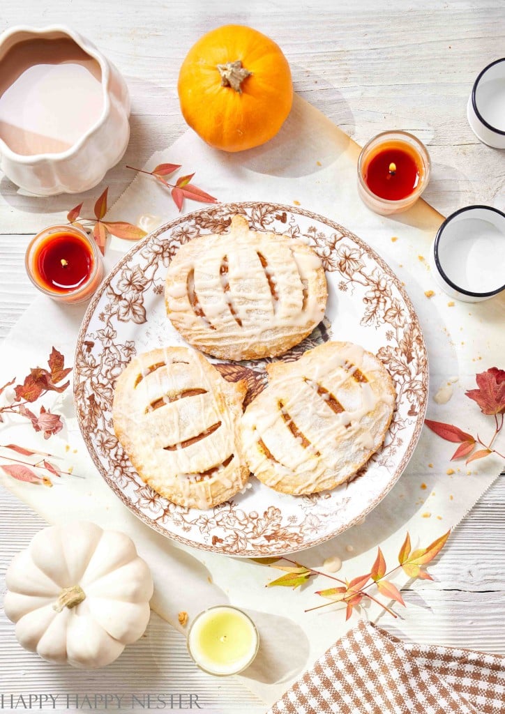 A decorative plate holds three glazed hand pies with slits on top, surrounded by autumn leaves, pumpkins, candles, and a pitcher on a light wooden table, perfect for serving as cozy fall or Halloween treats.
