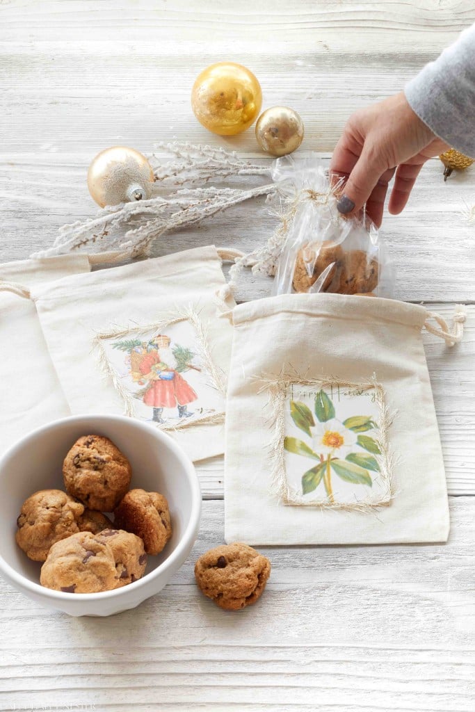 A hand places cookies in a clear bag inside a floral fabric gift bag—one of my favorite holiday home crafts. Nearby are a bowl of cookies, another festive gift bag, and gold ornaments on a white wooden table.