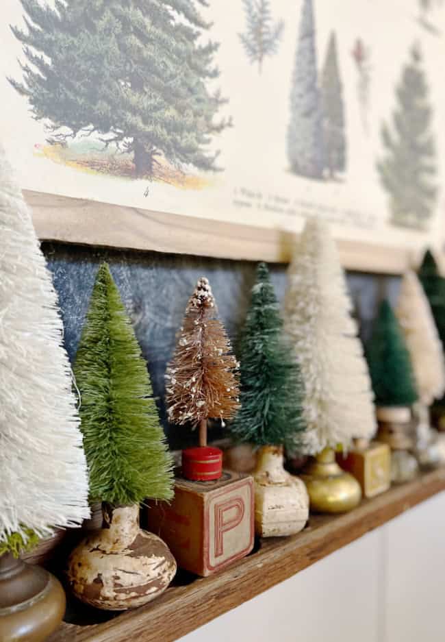 A row of small, vintage-style bottle brush Christmas trees—my favorite holiday home crafts—in various shades of green and white, each set in old wooden blocks and brass bases, sits on a shelf with a botanical tree print above.