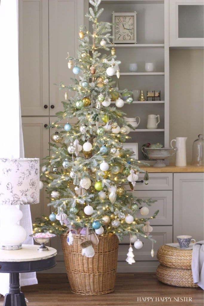 A decorated Christmas tree in a wicker basket stands in a cozy living room, adorned with vintage style Christmas ornaments and twinkling lights. Shelves with dishes, a clock, and a lamp are visible in the background.
