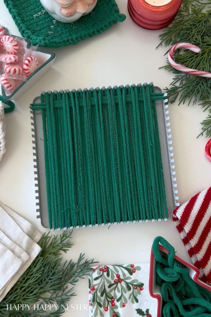 A green yarn potholder is woven on a square metal loom, surrounded by festive holiday items—perfect for DIY Christmas gifts—with a candy cane, peppermint candies, greenery, a red and white towel, and a Christmas-themed napkin.