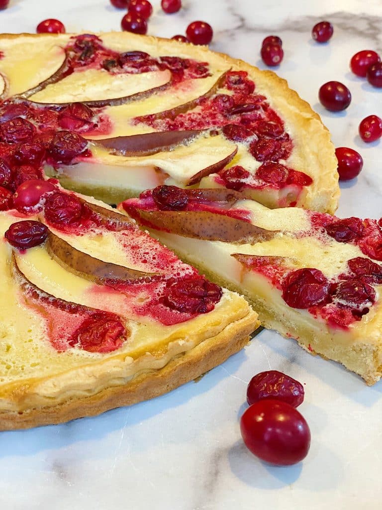 A sliced tart topped with fresh cranberries and pear slices sits on a marble surface, surrounded by scattered cranberries—the perfect Thanksgiving food with its golden crust and creamy filling.