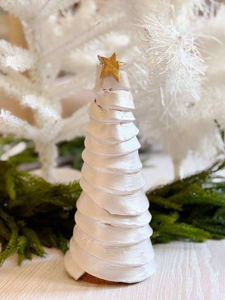 A white, spiral-shaped Christmas tree decoration with a gold star on top, set against white artificial trees and green foliage on a light surface—one of my favorite holiday home crafts.