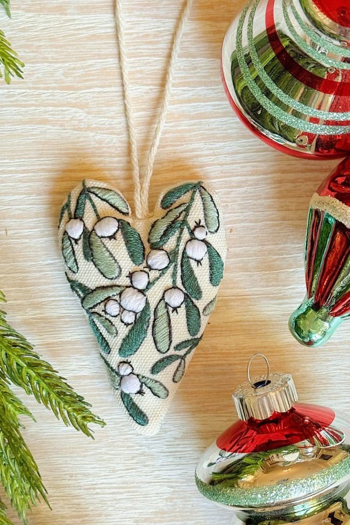 A fabric heart ornament embroidered with green leaves and white berries lies on a light wooden surface, surrounded by pine needles and shiny baubles—perfect for adding vintage style Christmas ornaments to your holiday décor.
