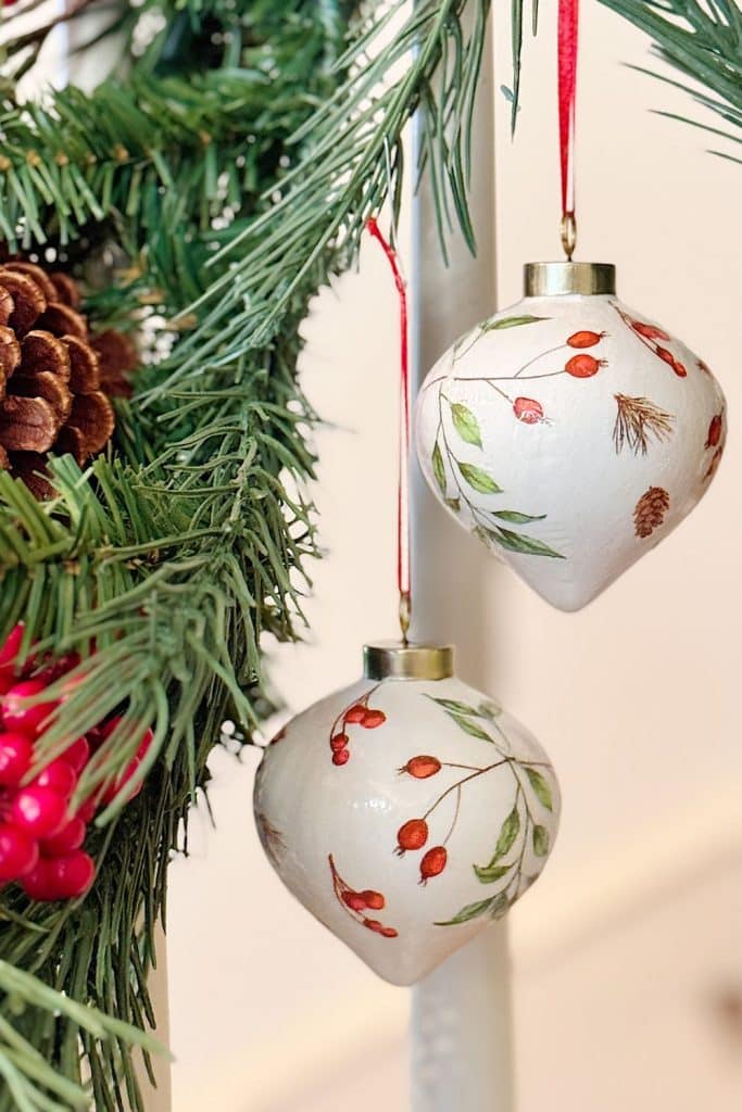 Two white Christmas ornaments with hand-painted red berries and green leaves hang from red ribbons on a pine garland, making festive gift ideas among pinecones and red berries.