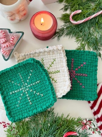 Three crocheted potholders—two green and one white—each with a stitched snowflake design sit on a festive table with greenery, a candle, peppermint candies, and more. These diy creations make charming potholder Christmas gifts for the holiday season.