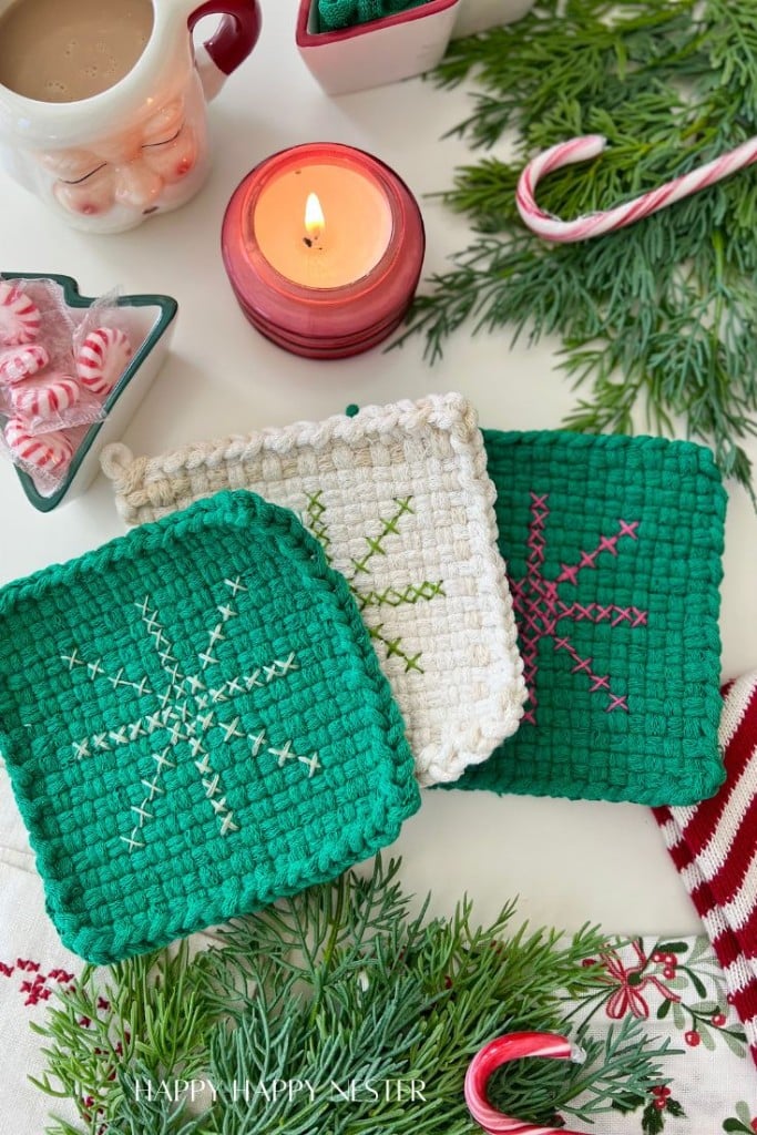 Three crocheted pot holders with stitched snowflake patterns sit on a festive table, showcasing my favorite holiday home crafts among evergreen branches, a lit candle, a mug, a heart dish of mints, and a candy cane for a cozy holiday scene.