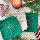 Three crocheted potholders—two green and one white—each with a stitched snowflake design sit on a festive table with greenery, a candle, peppermint candies, and more. These diy creations make charming potholder Christmas gifts for the holiday season.