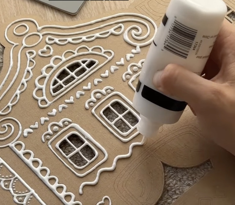 A hand squeezes white glue from a bottle, outlining and decorating a cardboard gingerbread house with intricate patterns and window details—one of my favorite holiday home crafts.