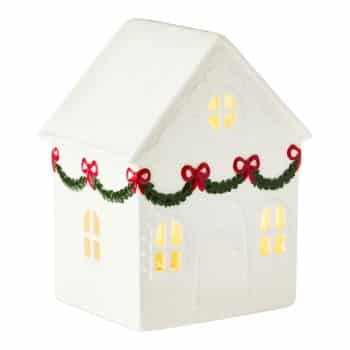 A white ceramic house with illuminated windows, decorated with green garlands and red bows along the roofline, evoking a festive holiday theme.