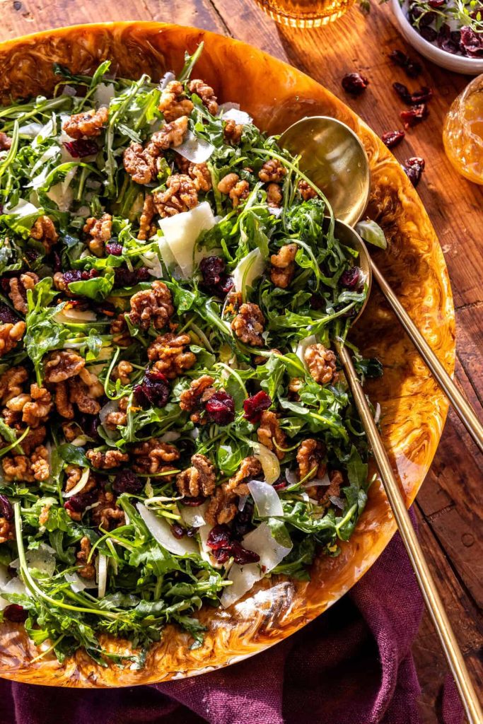 A large bowl of arugula salad topped with shaved parmesan, candied walnuts, and dried cranberries—perfect as a refreshing Thanksgiving food—rests on a wooden table with two serving spoons inside.
