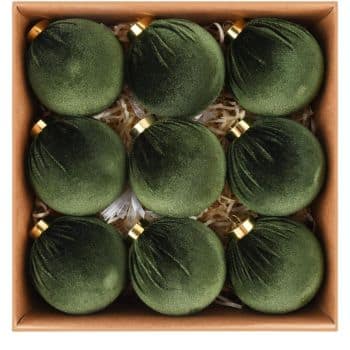 A box containing nine green velvet Christmas ornaments with gold caps, arranged in three rows on a bed of shredded brown paper.