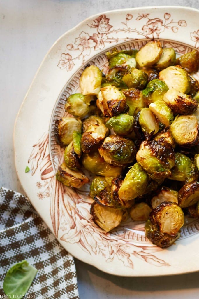 A vintage floral-patterned plate filled with roasted Brussels sprouts, golden and caramelized, sits on a light-colored surface next to a brown and white checkered cloth napkin—perfect as classic Thanksgiving food.