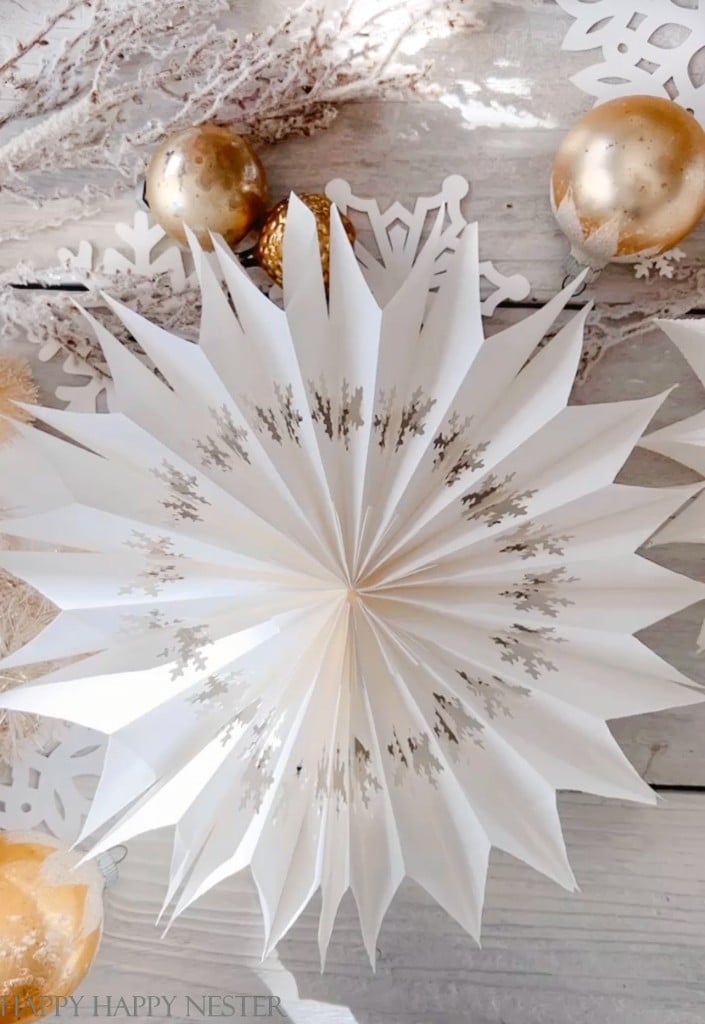 A white paper star decoration with silver snowflake patterns is displayed on a light wooden surface, surrounded by gold ornaments, white branches, and paper snowflakes—one of my favorite holiday home crafts.