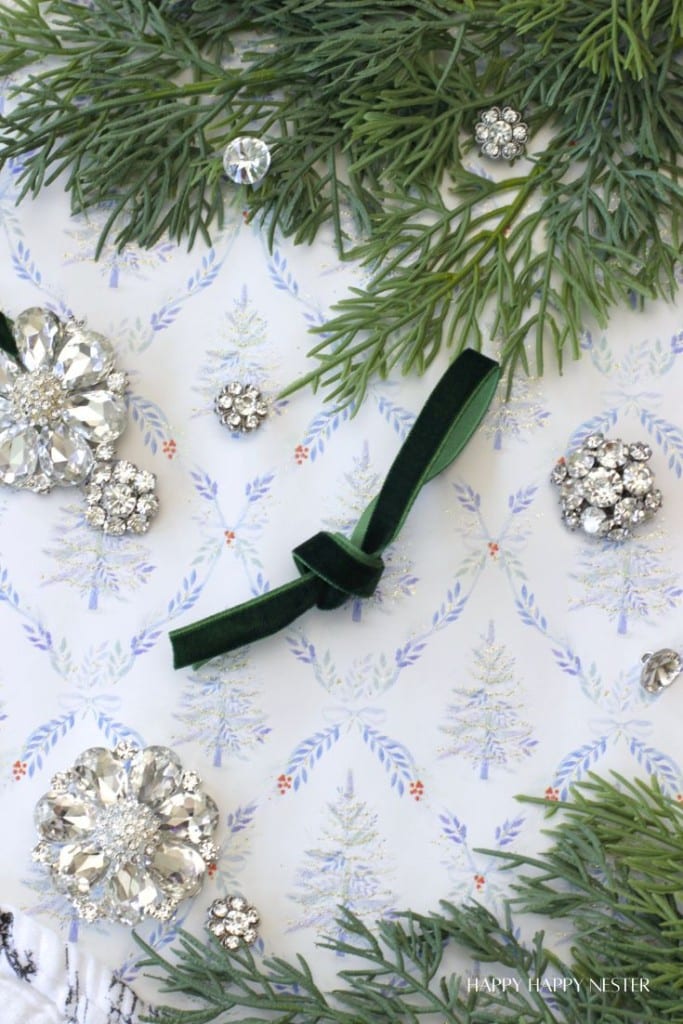 A festive arrangement with green pine branches, sparkling rhinestone brooches, small crystals, and a dark green velvet ribbon on decorative paper featuring a winter tree pattern.