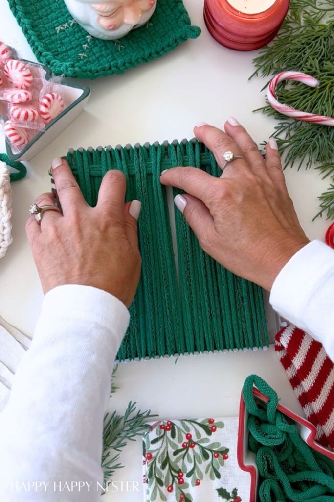 A person weaves green fabric loops on a loom, surrounded by holiday items like candy canes, greenery, peppermint candies, a Christmas-themed napkin, and a striped towel on a white surface.
