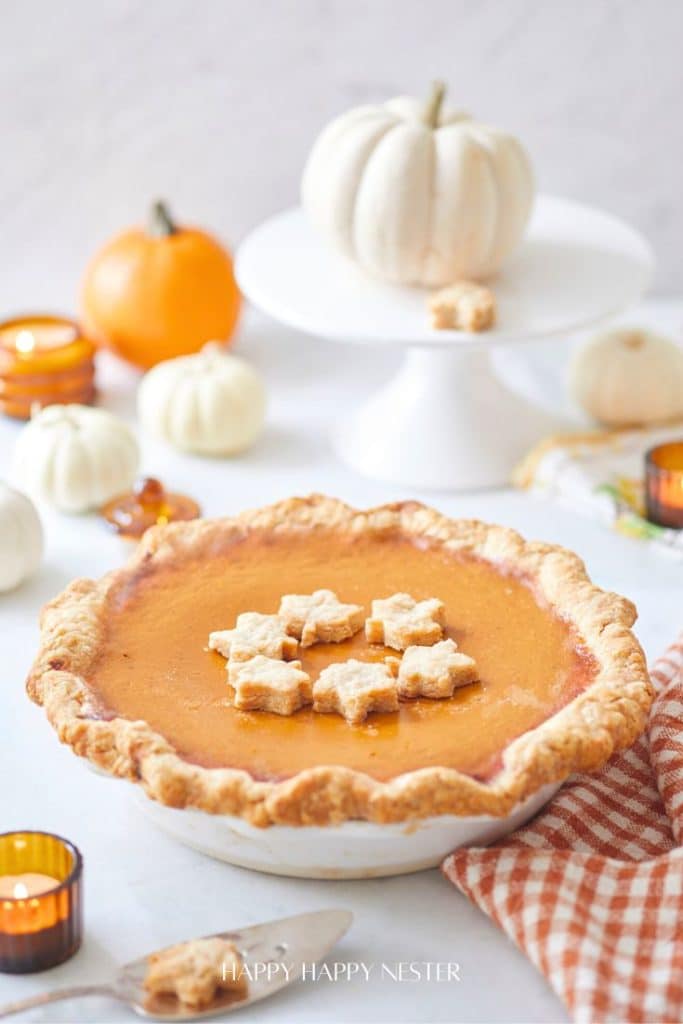 A pumpkin pie with a golden crust and star-shaped pastry decorations sits on a table, surrounded by thanksgiving food like small pumpkins, candles, and an orange-checked napkin. A white pumpkin is displayed on a cake stand in the background.