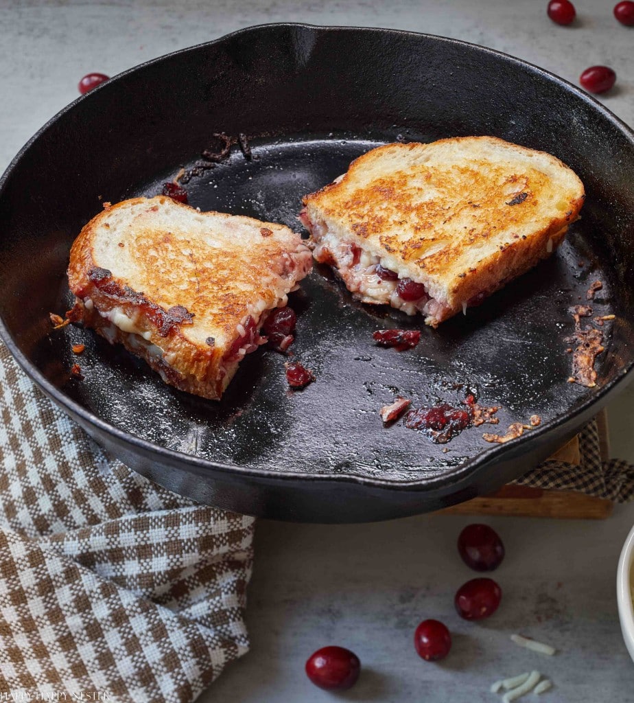 A grilled cheese sandwich with cranberries, a creative twist on classic thanksgiving food, is cut in half and sits in a black cast iron skillet. A checkered cloth and some loose cranberries are nearby on a light gray surface.