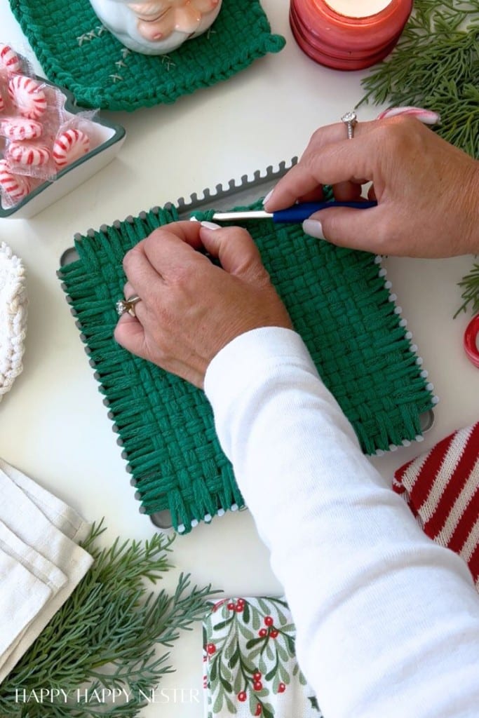A person uses a hook tool to weave green loops on a square loom, making a potholder. Festive decorations, peppermint candies, candles, and greenery are nearby on a white surface.