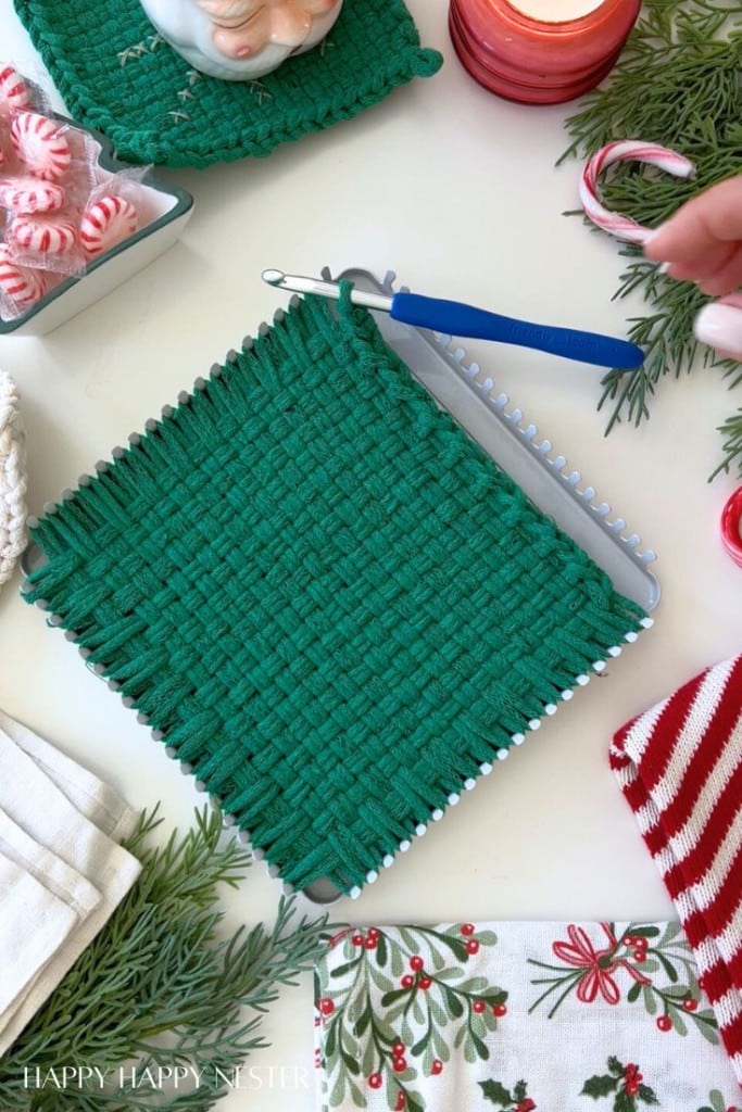 A green woven square potholder in progress on a loom with a blue hook, surrounded by Christmas decorations, candy canes, peppermint candies, greenery, and festive linens.