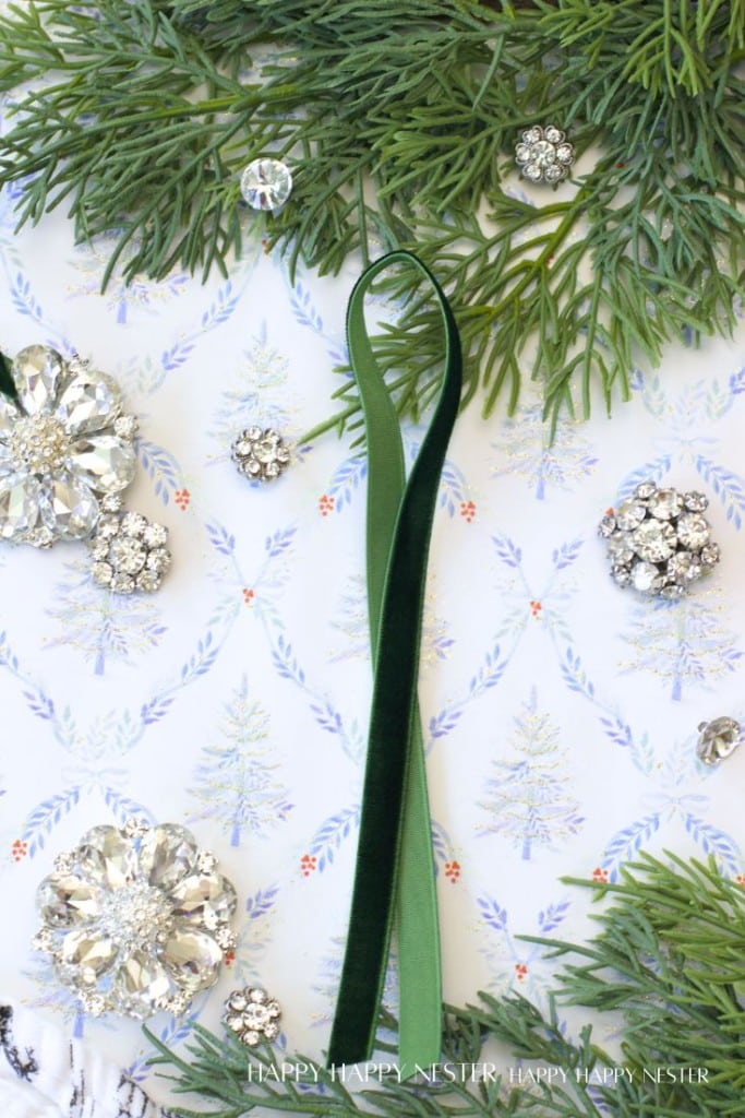 Sprigs of greenery, vintage rhinestone brooches, and a green velvet ribbon are arranged on patterned paper with small trees, evoking a festive, elegant holiday flat lay perfect for vintage style Christmas ornaments DIY inspiration.