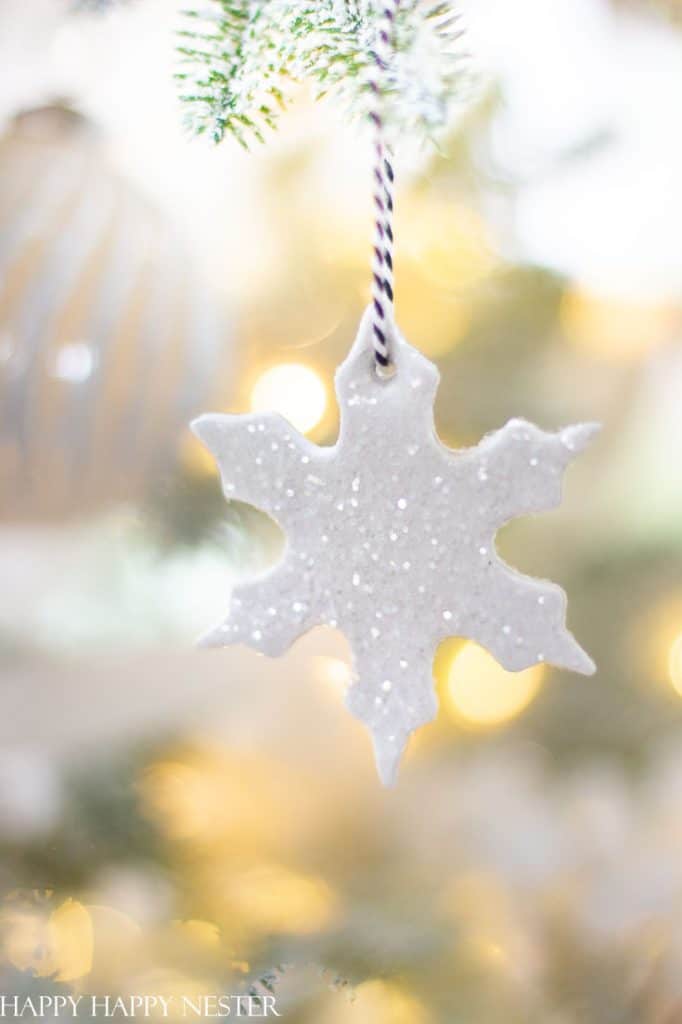 A white, glittery snowflake ornament—perfect for Christmas tree crafts—hangs from a black-and-white striped string on a Christmas tree branch, with blurred warm lights and soft greenery in the background.
