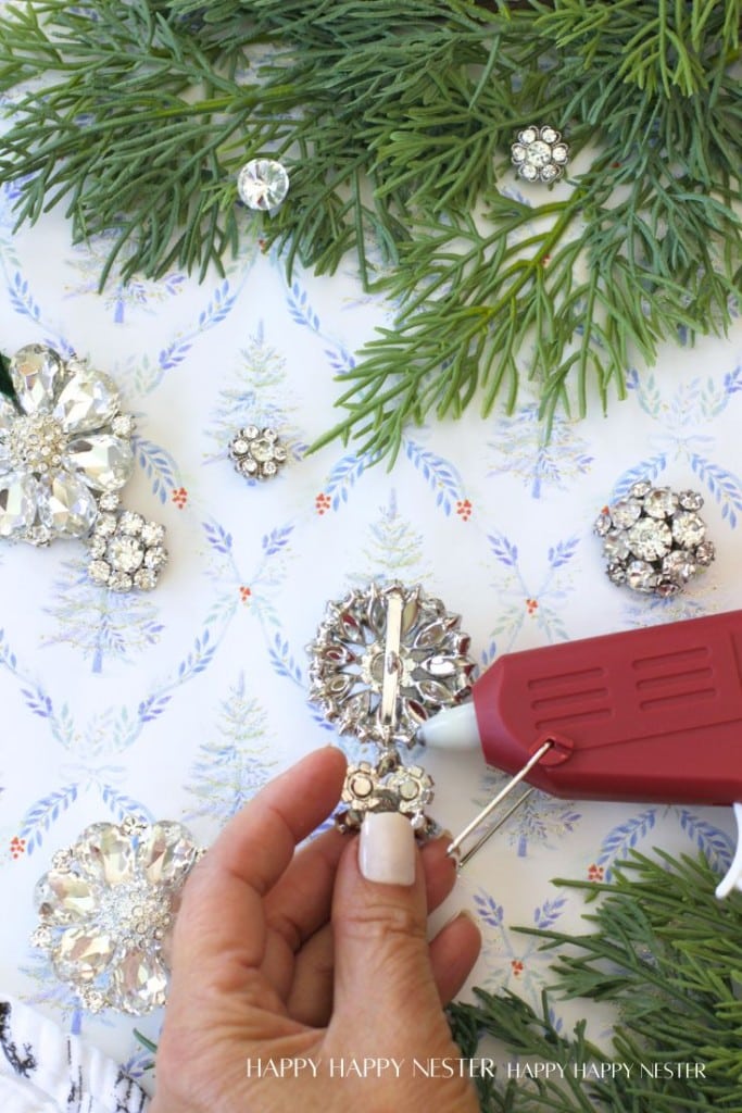 A hand holding a jeweled ornament near a red hot glue gun, surrounded by sparkling brooches on patterned paper and evergreen branches.