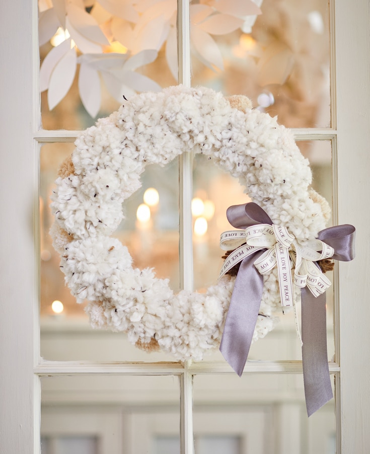 A fluffy white wreath, one of my favorite holiday home crafts, decorated with a gray and white ribbon bow, hangs on a glass pane door. Soft lighting and white paper leaves in the background create a cozy, festive atmosphere.