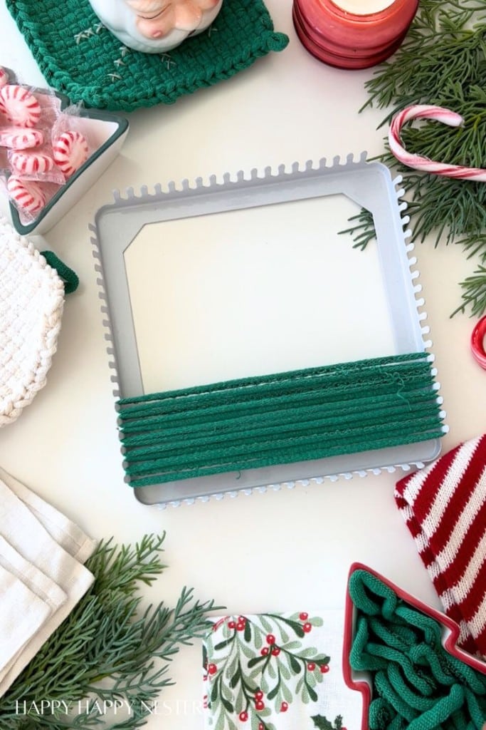 A loom with green yarn partly woven on it sits on a festive table surrounded by Christmas decorations, greenery, candy, and holiday-themed textiles in red, white, and green.