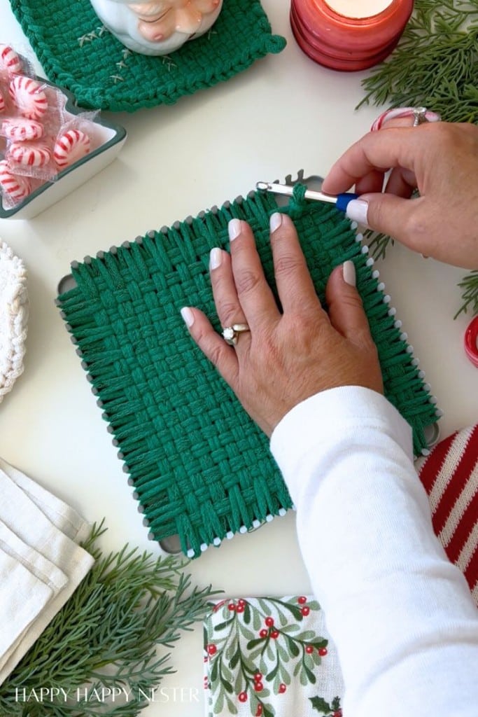 A person’s hands weaving a green potholder on a loom, surrounded by festive holiday decor, greenery, candy, and candles—perfect for diy Christmas gifts on a white table.