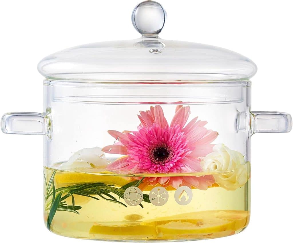 A clear glass Simmer Pot with handles and a lid, filled with liquid, lemon slices, rosemary, a pink flower, and white flowers. Icons are printed on the pot—perfect for experimenting with new Simmer Pot Recipes.