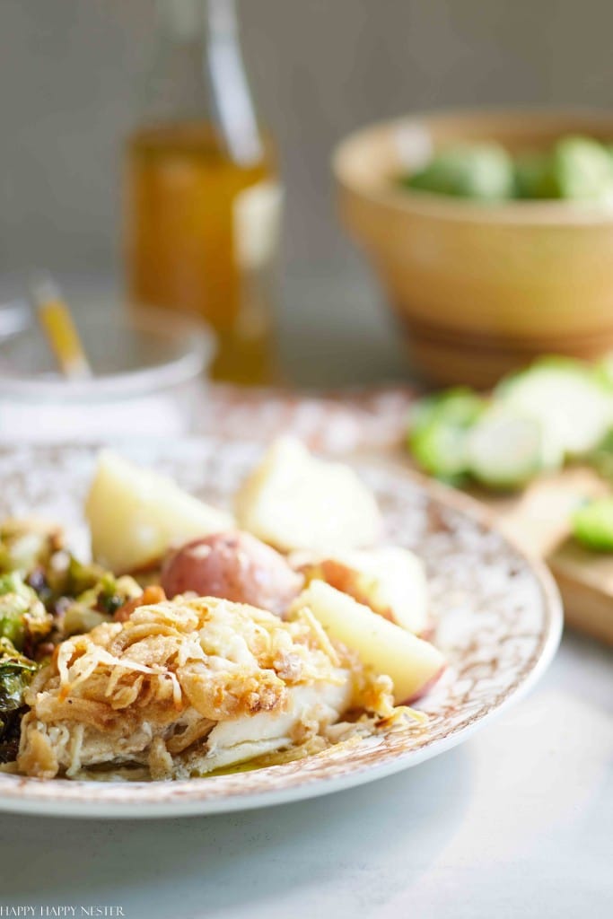 A plate with baked fish topped with onions, boiled baby potatoes, and roasted vegetables sits on a table—a fresh take on Thanksgiving food. A bottle of olive oil and a bowl of Brussels sprouts are blurred in the background.