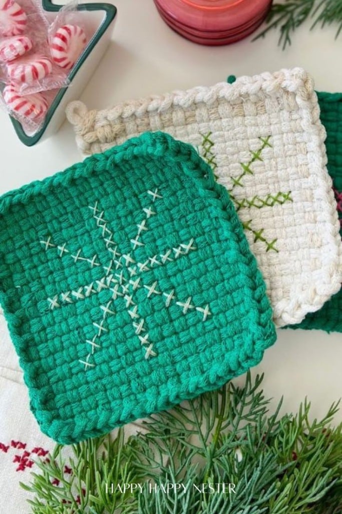 Two square, handwoven coasters with cross-stitched snowflake designs in green and white yarn are displayed on a festive table with pine branches and peppermint candies.